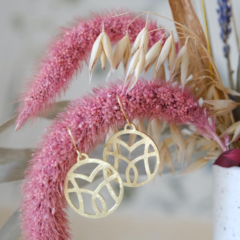 Brincos grandes de ouro reciclado.O motivo VM forma subtilmente um lótus. Brincos pendurados numa flor cor-de-rosa seca.