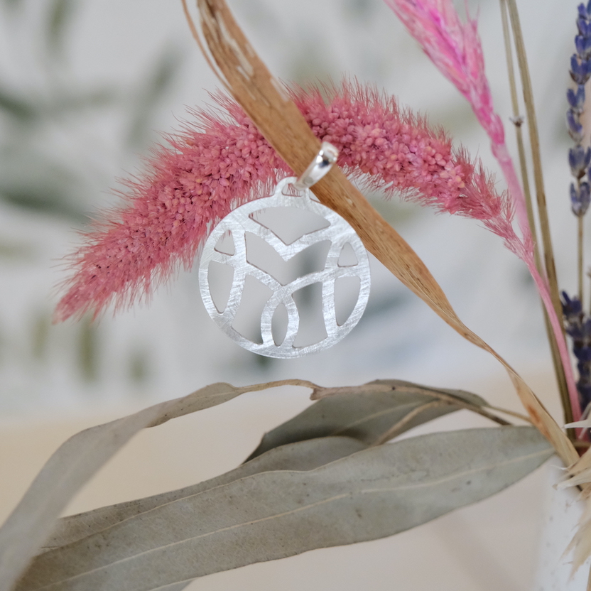Medalha em prata reciclada. O motivo VM forma subtilmente um lótus. A medalha pende de uma flor rosa seca.
